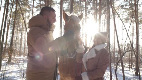 happy couple caressing horse and chatting in forest on winter day