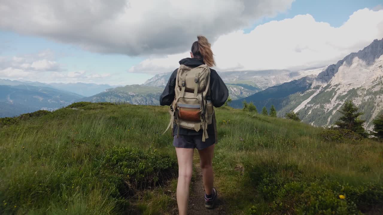 Sırtında çantasıyla, arka planda Dolomitler varken dağ yolunda yürüyüş yapan bir doğa yürüyüşçüsü, turist kadın gezginin arkadan görünümü