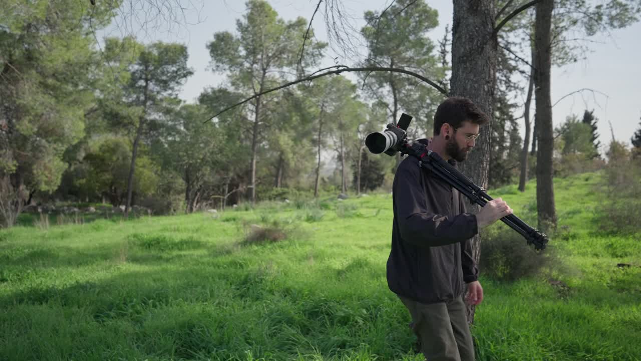 Pembuat konten berjalan perlahan melalui hutan sambil membawa kamera dan tripod selama pemotretan di luar ruangan.