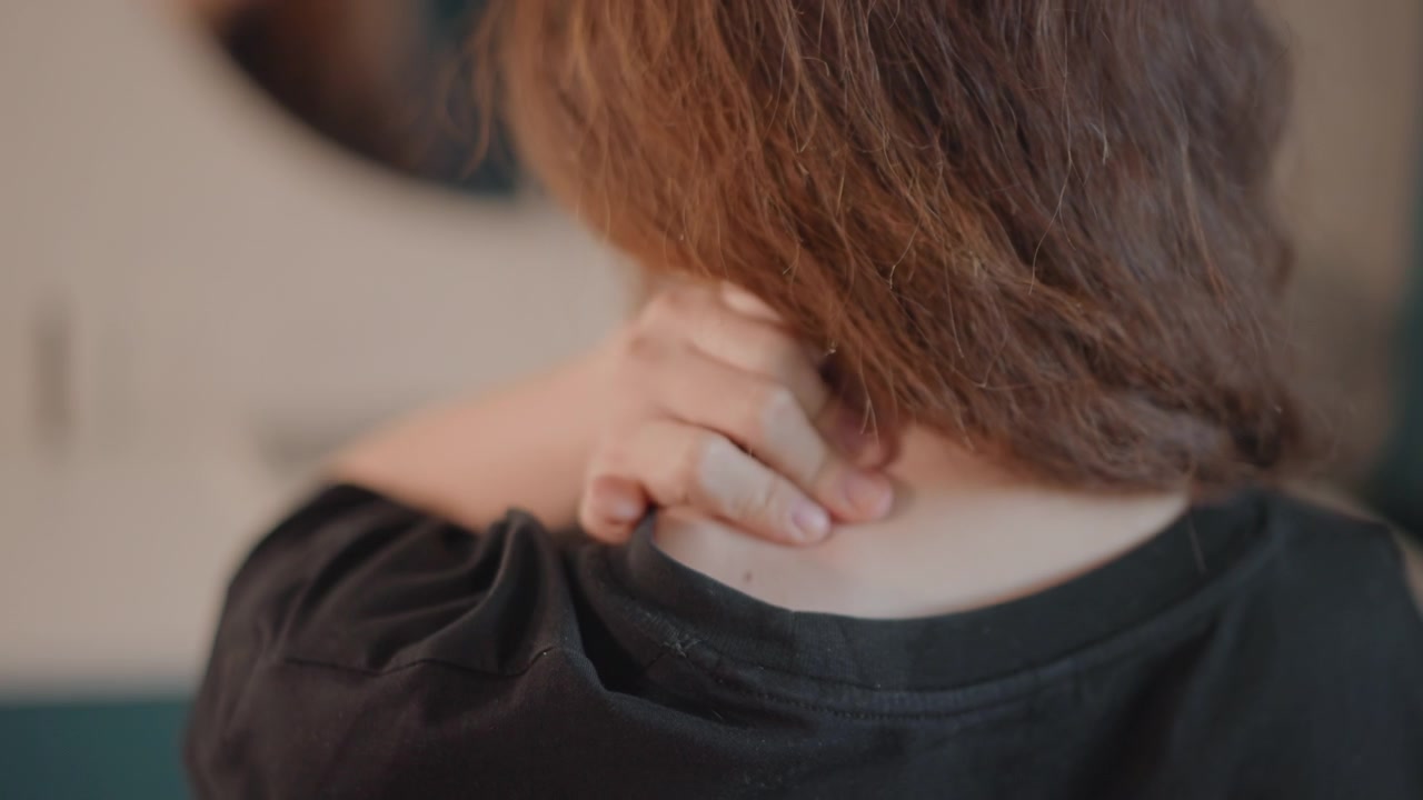 Primo piano posteriore di un adulto che si protende sopra la spalla per premere i polpastrelli nella parte superiore della schiena e alla base del collo, con una leggera inclinazione in avanti e il contatto della mano che suggerisce auto-massaggio, rilascio della tensione e tranquillità