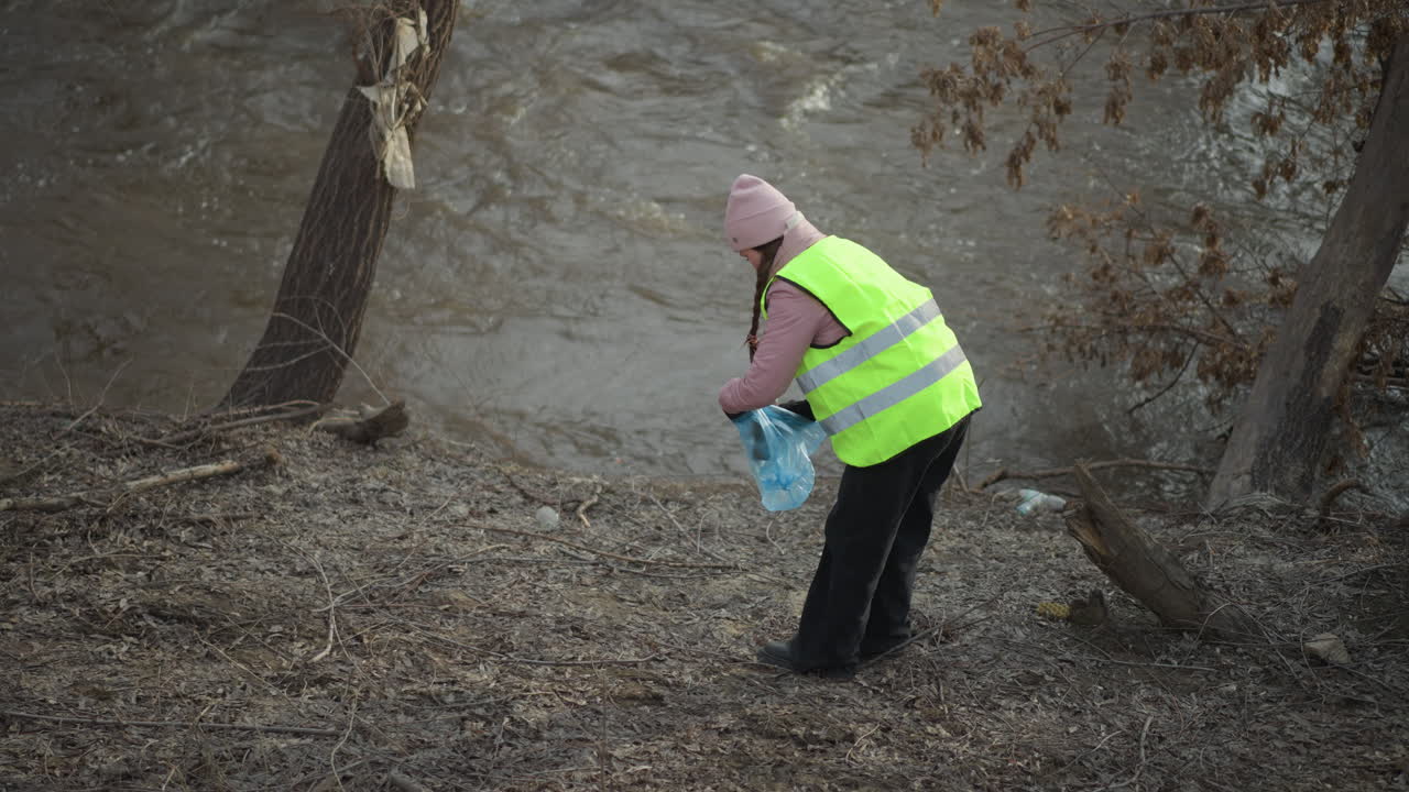 Woman in bright safety vest collects trash along muddy riverbank during cleanup, holding blue plastic bag, wearing pink beanie and gloves, engaging in environmental volunteer work near flowing water