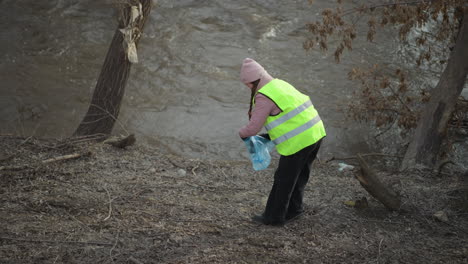 woman in bright safety vest collects trash along muddy riverbank during cleanup, holding blue plastic bag, wearing pink beanie and gloves, engaging in environmental volunteer work near flowing water