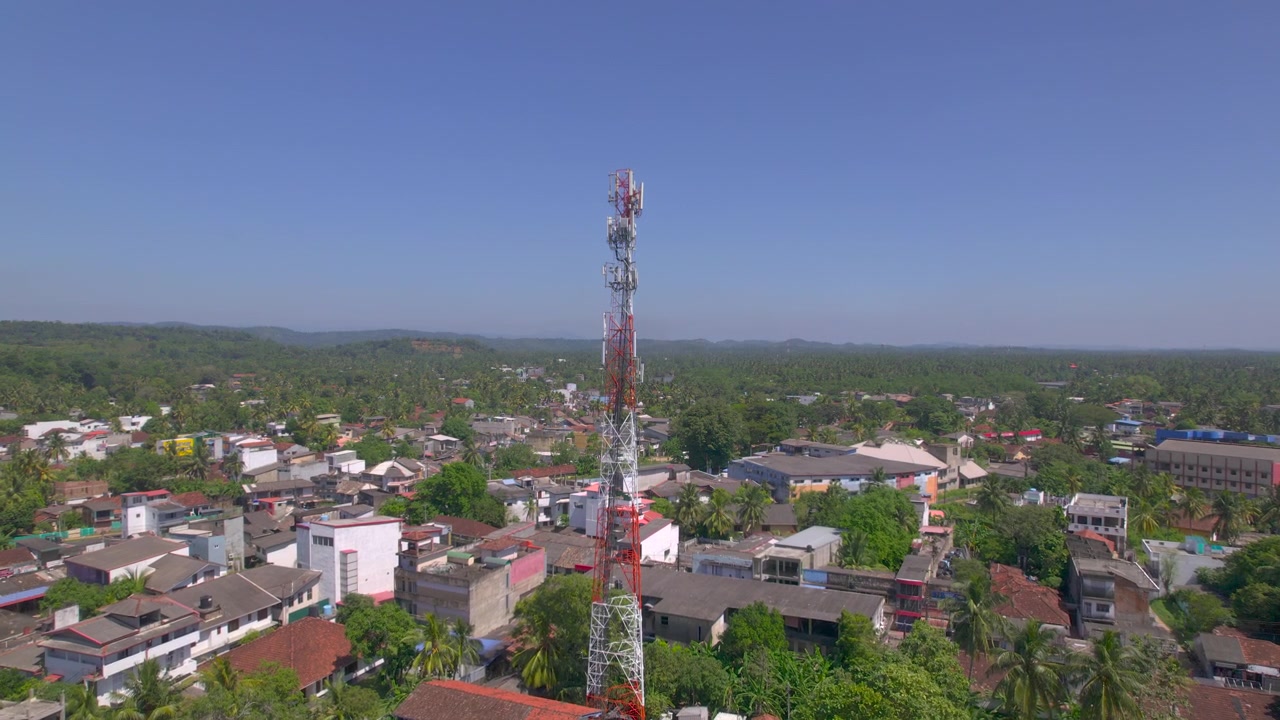 Droneflyvning afslører et højt telekommunikationstårn, der rejser sig over en kompakt srilankansk by med lave huse, røde tage, rigelige palmer og grønt tropisk landskab, der strækker sig til horisonten nedenfor.