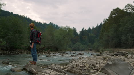 Man-standing-on-rocky-shore-of-river.-Hiker-touching-water-in-river-with-hand