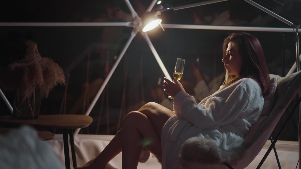 mujer contemplativa transfiere vidrio entre las manos mientras está sentada en una silla cómoda. cómoda en la luz tenue envuelve a mujer pensativa sentada en túnica acogedora