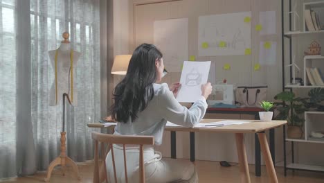 아시아 여성 디자이너의 뒷면은 손 가방을 보면서 머리를 긍정적으로 흔들고 손으로 레이아웃을 스케치합니다.