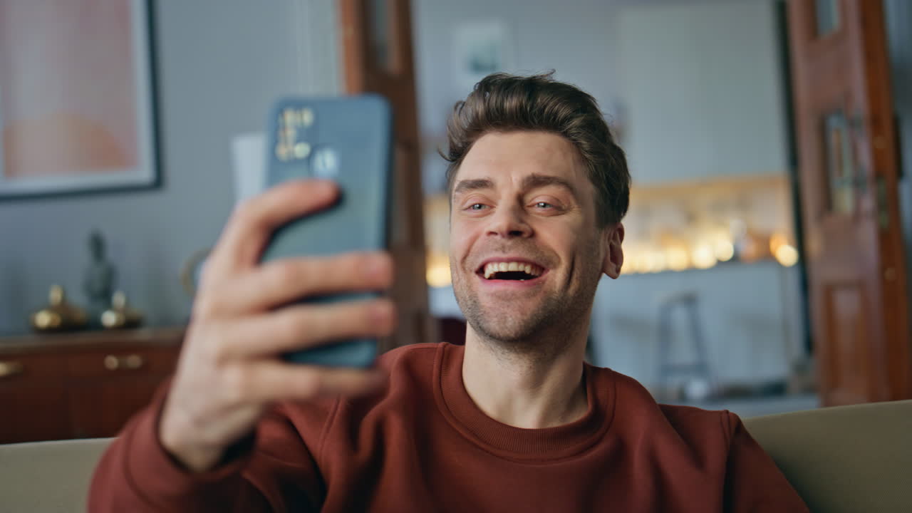 Smiling hipster video calling by mobile phone on couch close up. Man talking