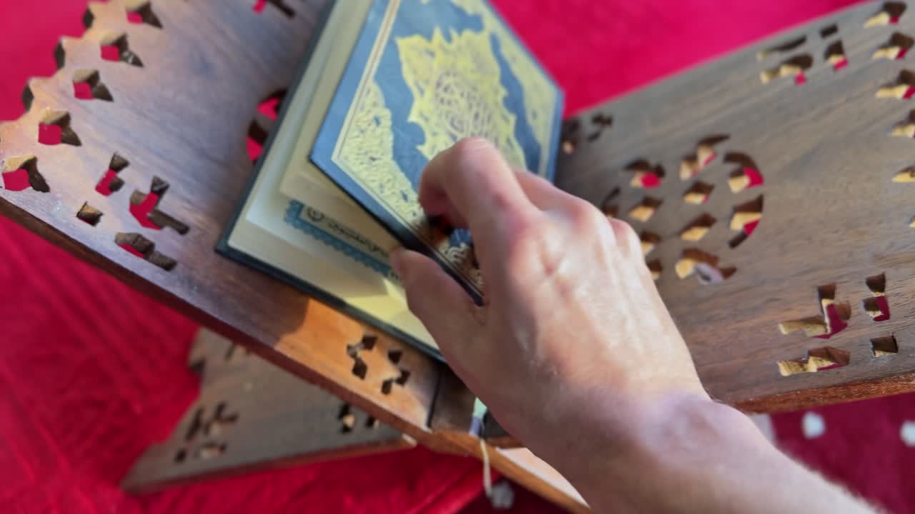 Close-up in 4K of a hand flipping through the Quran placed on a wooden rehal over a red prayer rug. Ideal for Islamic content, education, Ramadan visuals, and spirituality projects.