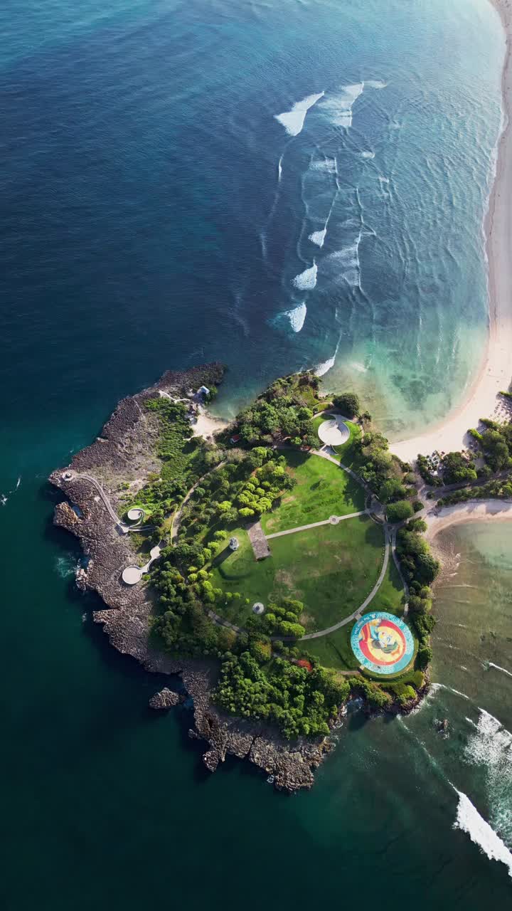 Vertical drone view of a rocky peninsula with deep blue ocean, white sand beach and coastal park, volcanic rocks and clean surf lines shaping a bold scenic shoreline