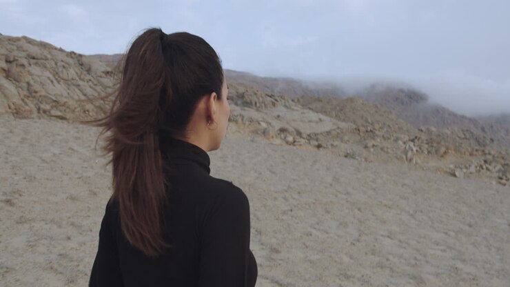 Close-up tracking of a beautiful woman from behind dressed in black walking through a mysterious desert while the wind blows in the afternoon