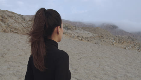 close-up tracking of a beautiful woman from behind dressed in black walking through a mysterious desert while the wind blows in the afternoon