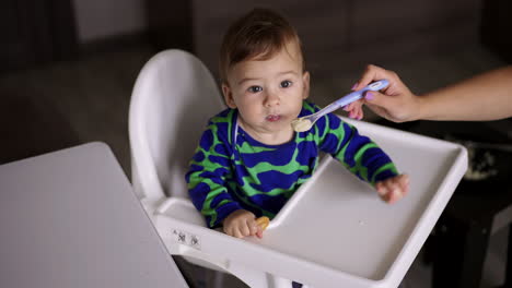sweet kid nutrition from a spoon. lovely boy sitting at the feeding table is fed by his mom. high angle view. blurred backdrop.