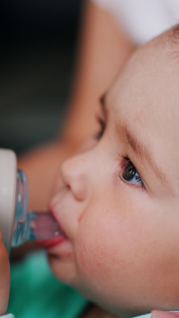 Lovely Caucasian child drinking water from a bottle and smiling. Mommy's hand hold a bottle for her son. Close up. Vertical video
