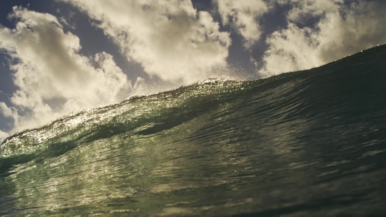 Sunlit ocean wave crest glowing beneath bright clouds and sky in smooth slow motion