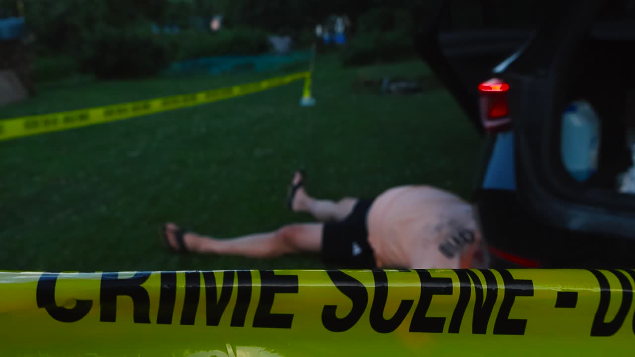 Police forensics yellow colored crime scene tape ribbon pulled infront of a crashed car with a male victim laying on the ground next to it at summer evening while the car lights are blinking slowly