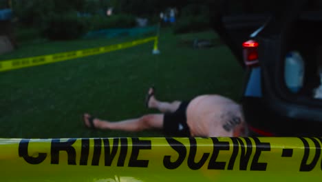 police forensics yellow colored crime scene tape ribbon pulled infront of a crashed car with a male victim laying on the ground next to it at summer evening while the car lights are blinking slowly