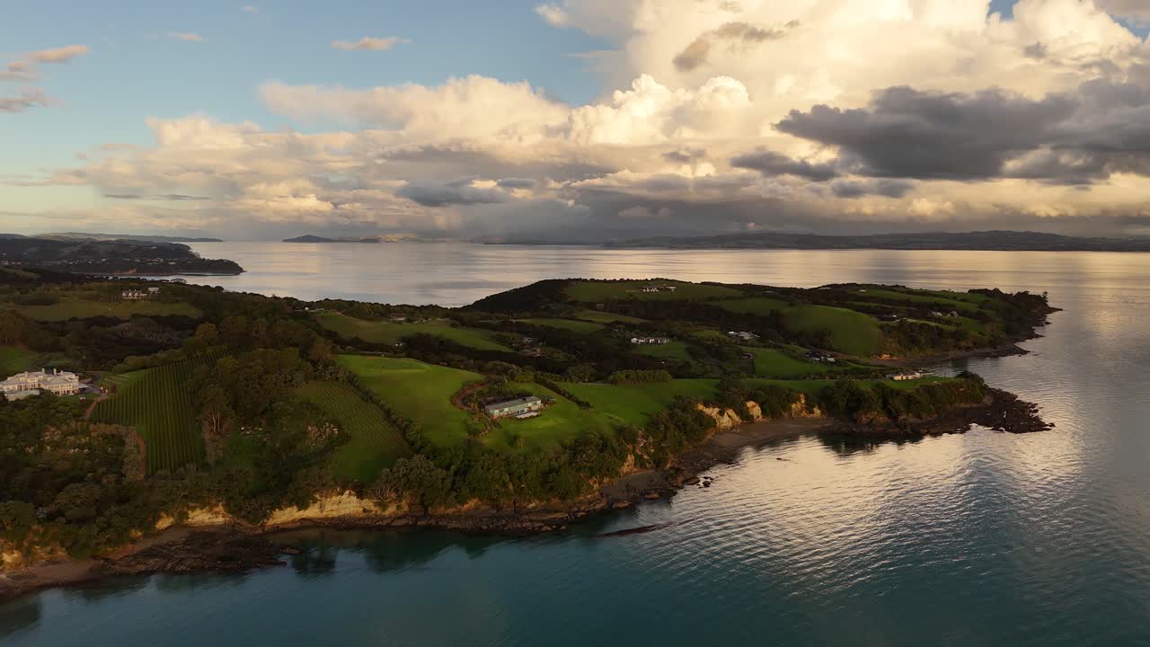 Aerial wide shot of green idyllic landscape with cliffs of Waiheke Island, New Zealand. Golden Sunset with clouds at sky.- Tranquil water bay with luxury villas.