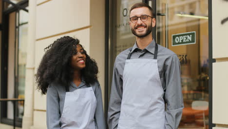 barista caucásico y barista afroamericana de pie en la puerta de un café moderno, sonriendo y mirando la cámara