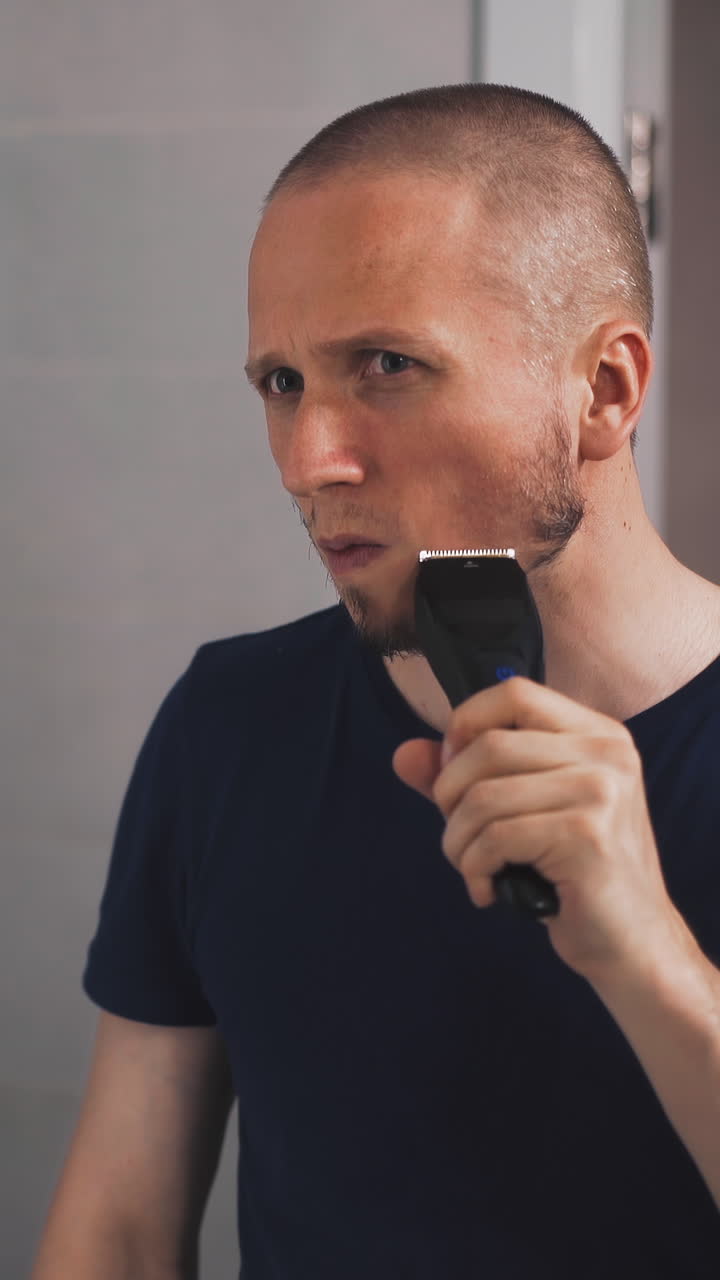 concentrated young man in black t-shirt shaves beard with modern electrical razor looking in large mirror in light bathroom at quarantine closeup