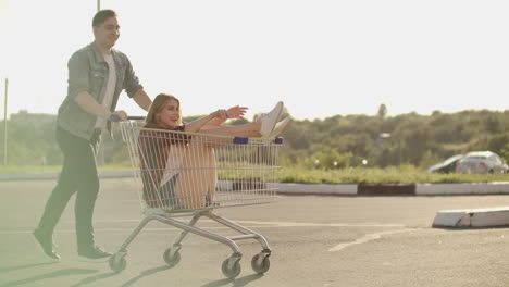 side view of a young man and woman having fun outdoors on shopping trolleys. multiethnic young people racing on shopping carts. on the parking zone with their