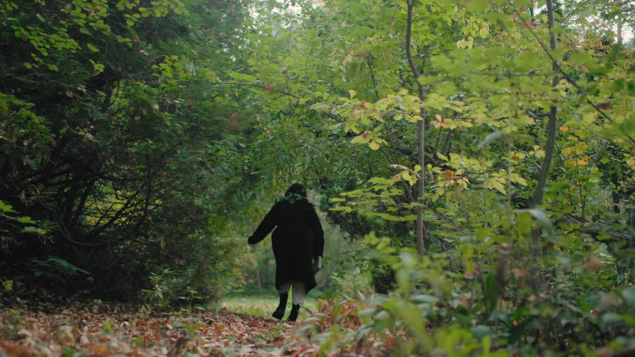 Back view of pensive woman in black coat walking into forest path with book in hand, surrounded by fresh green leaves, slow solitary stroll toward deeper woods, reflective escape in nature