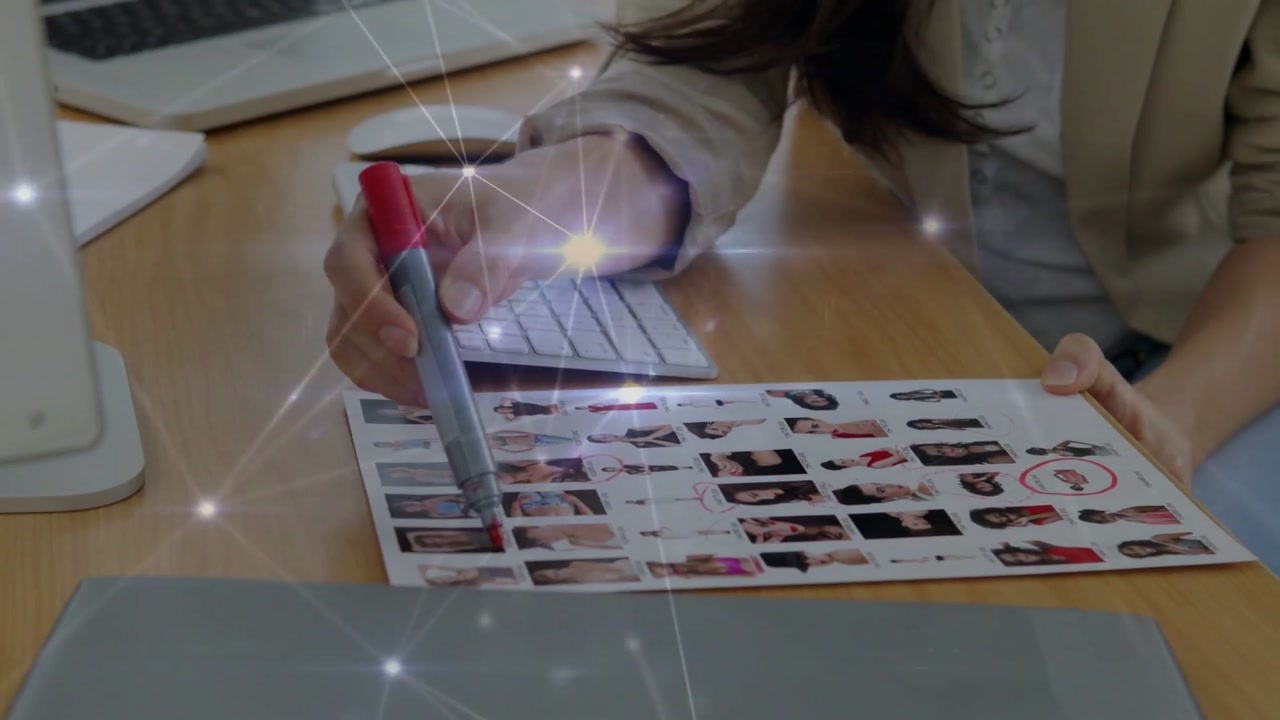 Woman placing thumbnail sheet by keyboard, picking up red marker, circling portraits for casting