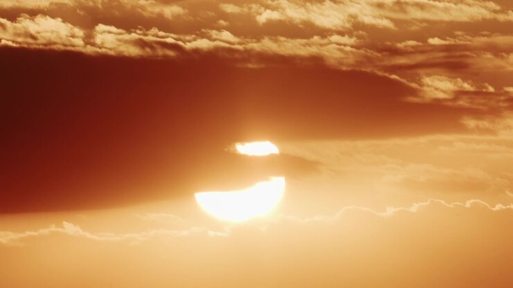 Wide shot of the sun slowly lowering behind a dense band of dark clouds, with a glowing horizon line beneath