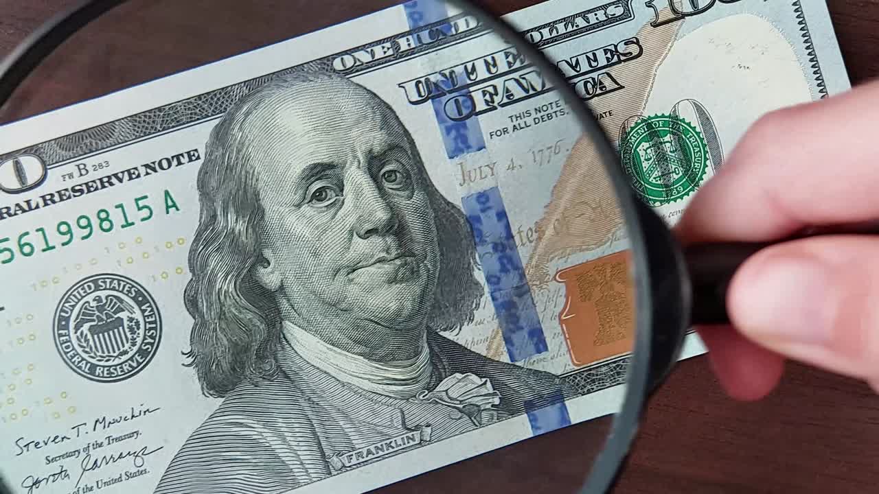 una mano con una lupa examina un billete de cien dólares. el concepto de dinero falso, finanzas, economía