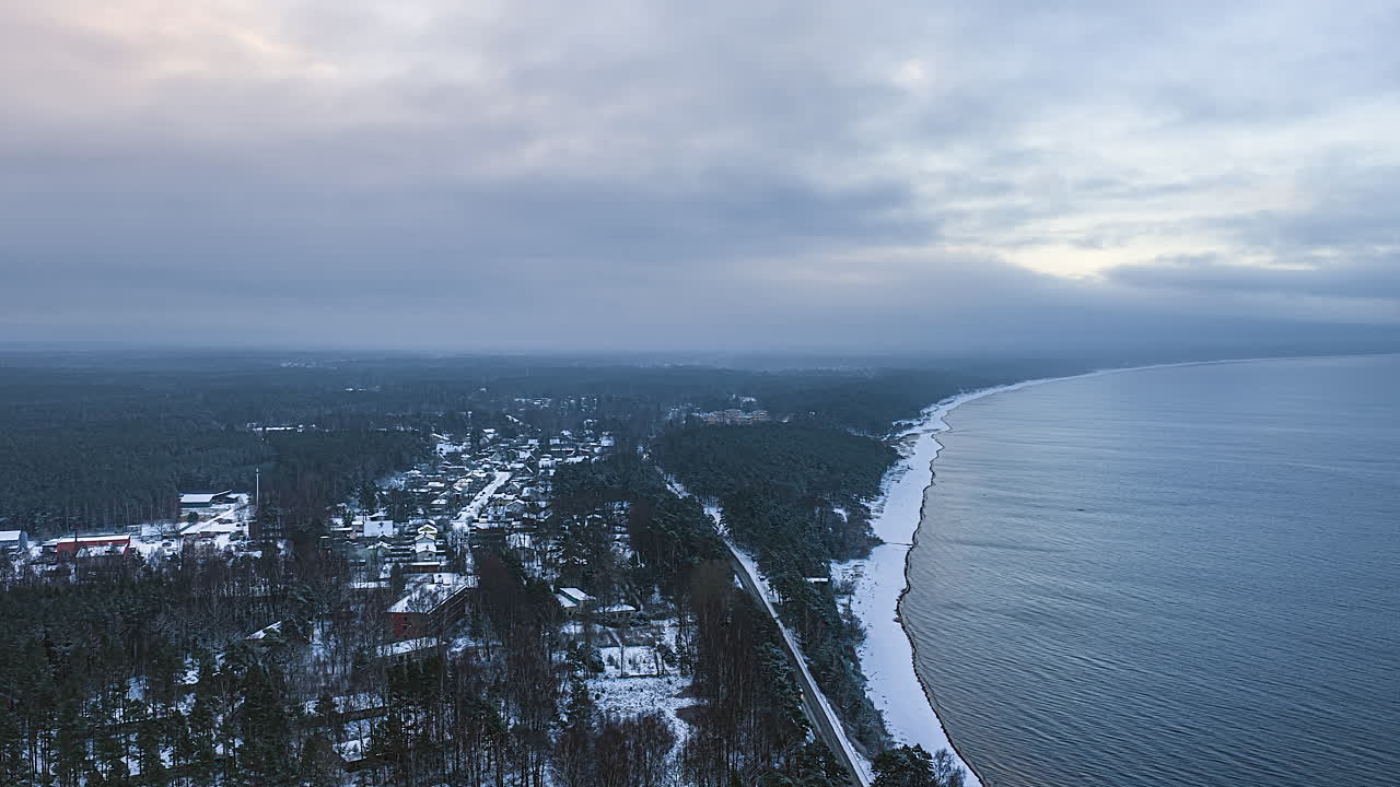 Drone-timelapse over en snødekt kystby og furuskog i skumringen, med varmt lys som bryter gjennom skyene over et rolig hav og en lang vinterkystlinje