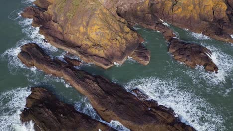 A-mesmerizing-aerial-shot-begins-at-the-ocean-cliffs,-capturing-the-power-of-crashing-waves,-then-pans-upward-to-reveal-Dunnottar-Castle-and-the-scenic-fields-beyond