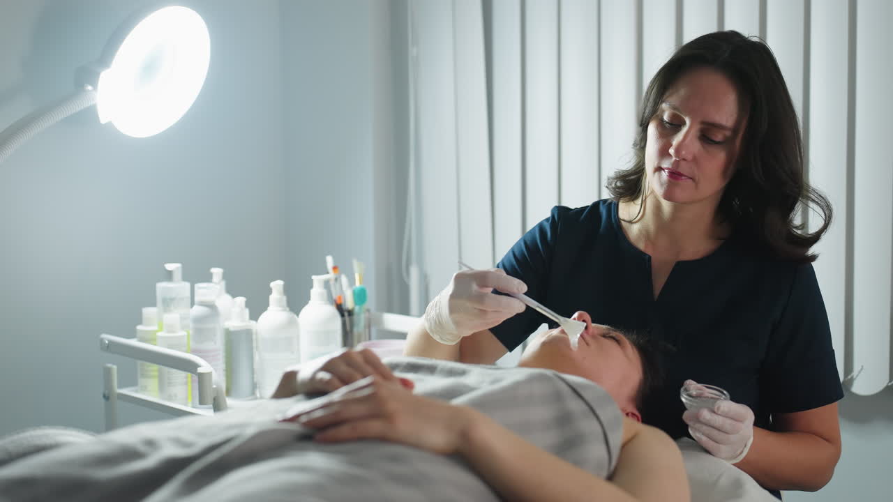 Wellness technician in gloves dips brush into clear gel mixture then gently applies cleansing mask onto closed eyes and cheeks of relaxed client lying on massage bed under bright lamp