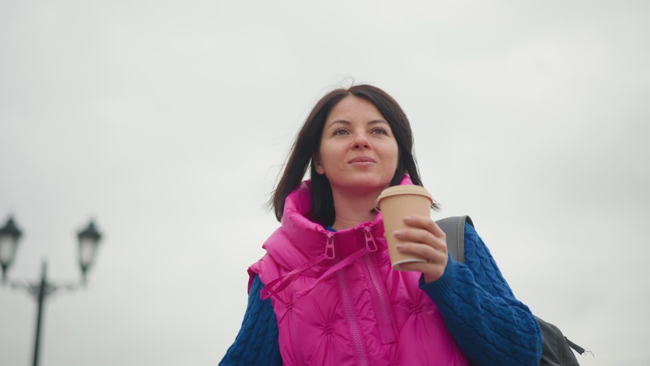 Donna che tiene in mano una tazza di caffè all'aperto, cielo nuvoloso, lampioni sfocati, passeggiata in città con gilet imbottito rosa e maglione blu, zaino in spalla, beve un sorso da una tazza da asporto, aria contemplativa