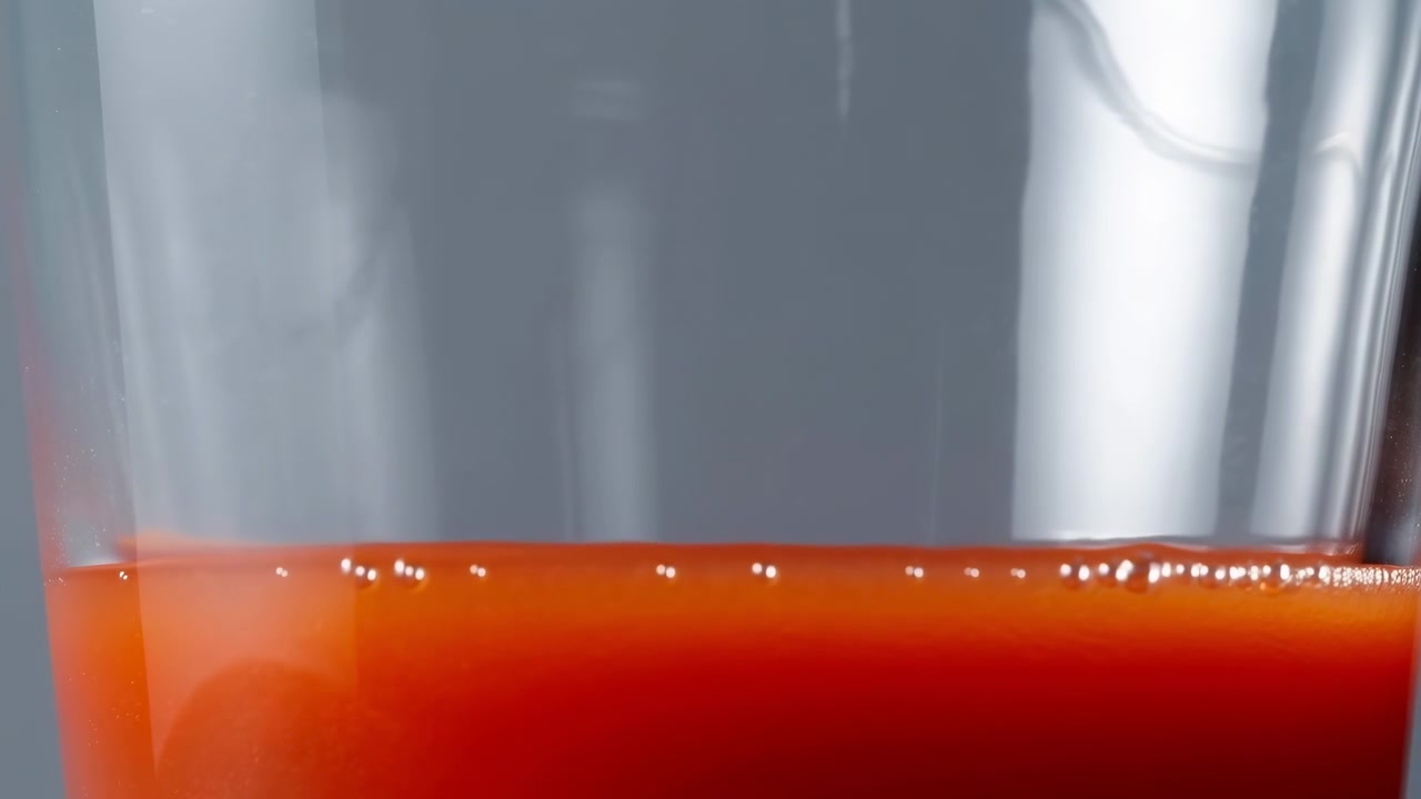 Primo piano di un contenitore di vetro trasparente che contiene un liquido rosso-arancio brillante con piccole bolle vicino alla superficie, ambientato su uno sfondo grigio delicatamente sfocato