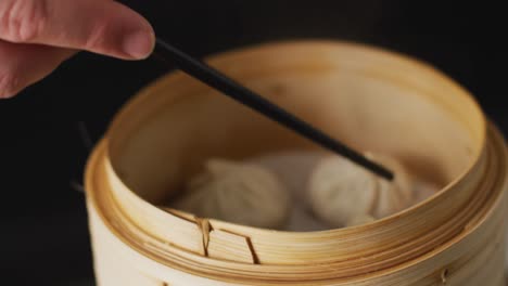 hand holding chopsticks with dim sum dumpling in bamboo steamer on black background