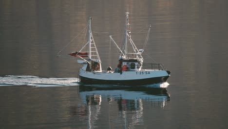 a small fishing ship moving slowly in the still fjord waters leaving the water trail in its' wake