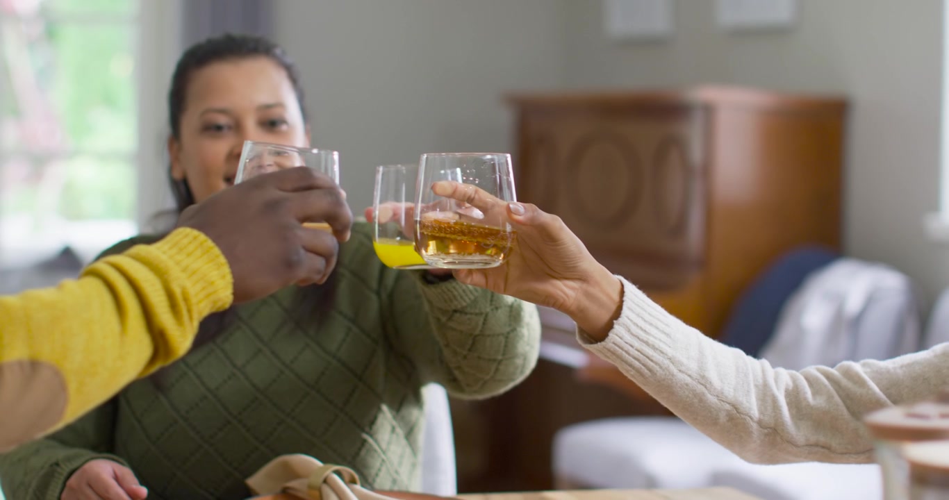 Friends raising short glasses at dining table, woman in green sweater clinking for meal toast