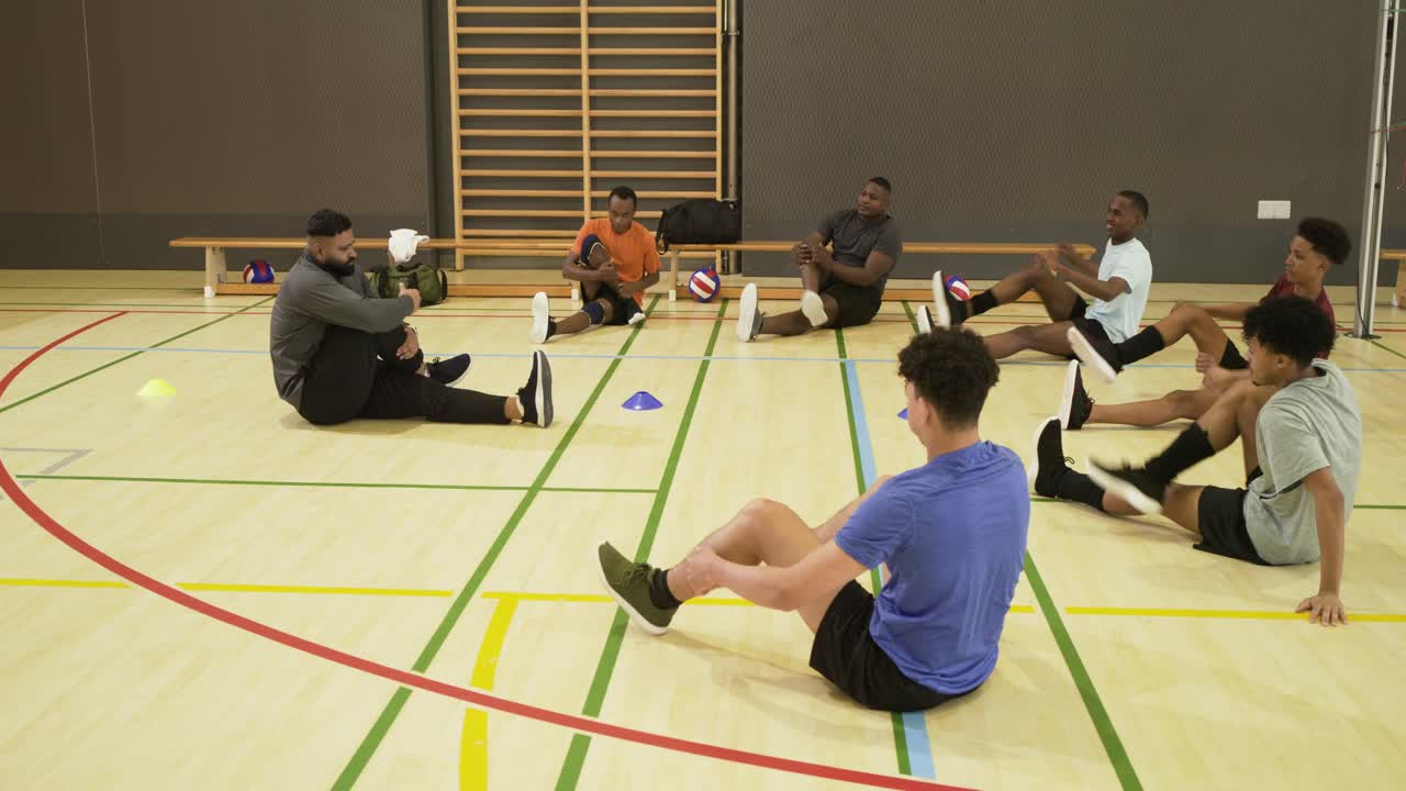 Diverses Männerteam folgt Anweisungen des Trainers, führt sitzende Dehnübungen zum Aufwärmen im Fitnessstudio mit Hütchen durch