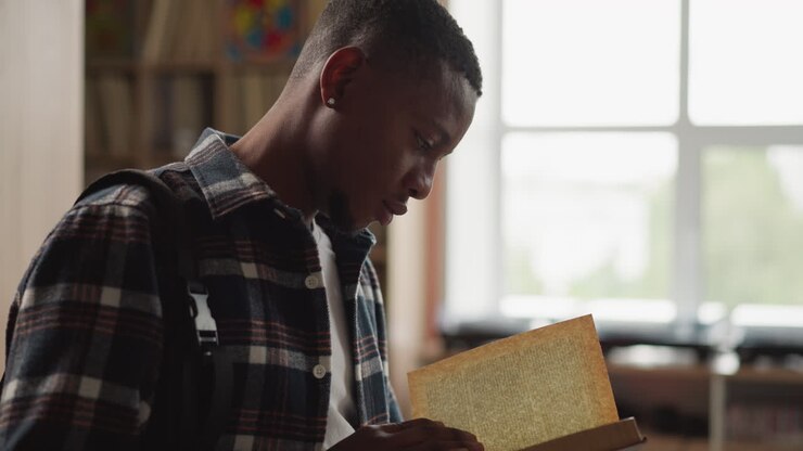 Man reads book in university library. Young African American man interested in scientific article in textbook in information center. Reader holds novel