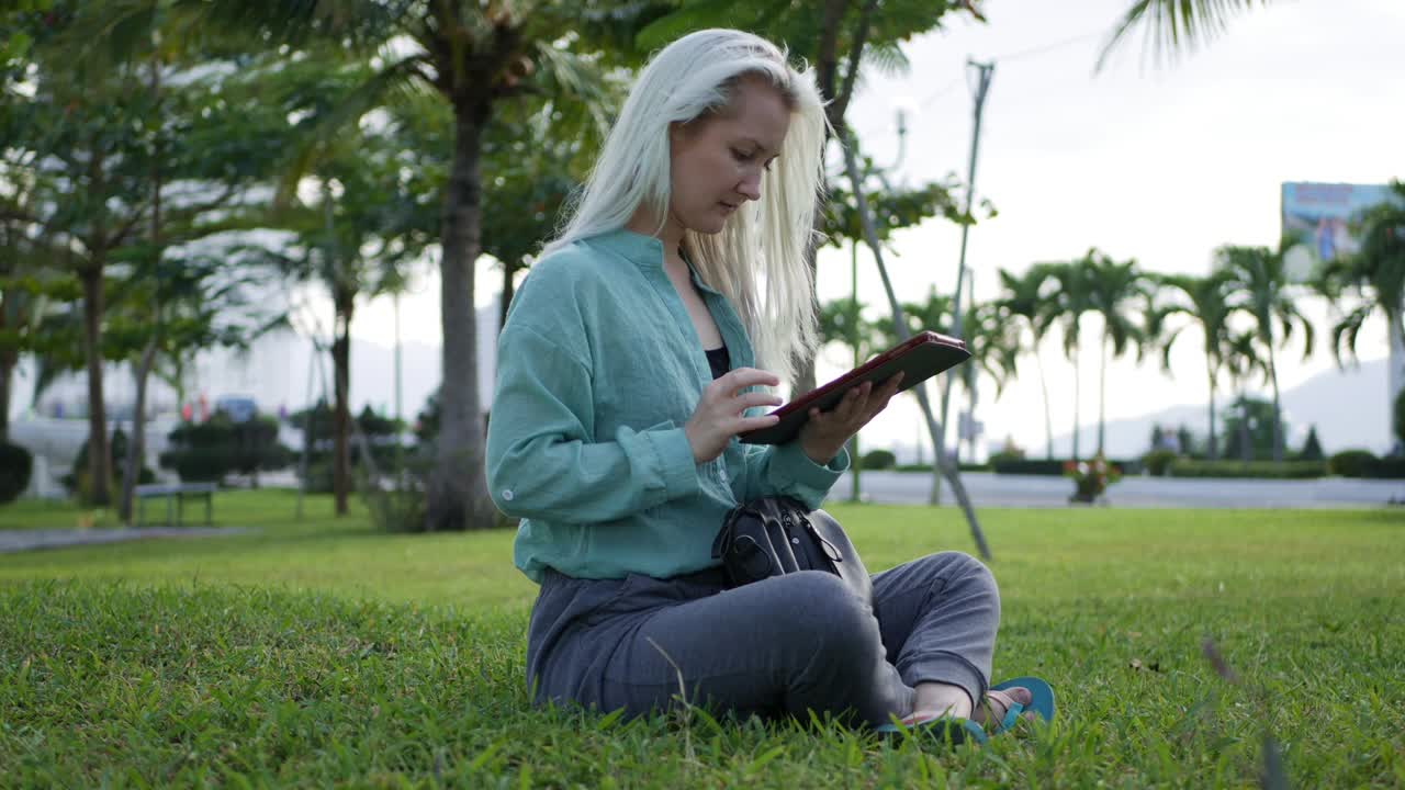 hermosa mujer delgada con el cabello largo rubio en camisa verde se sienta en el suelo y el uso de teléfono inteligente sobre el fondo del parque. niña en la pantalla cuadrada tocando y sonriendo
