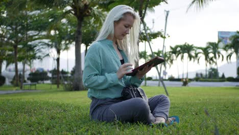 beautiful slim woman with long blonde hair in green shirt sits on the ground and using smartphone over background the park. girl on the square touching screen and smile