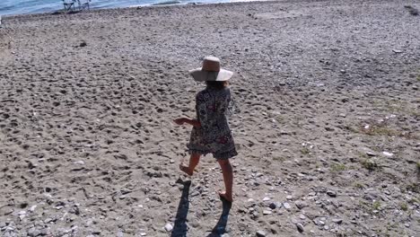 Aerial-flight:-Young-woman-in-straw-hat-walks-on-the-beach-towards-the-sea,-Turkey