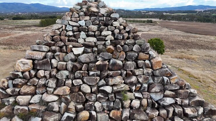 Dronekamera stiger op over en stor stenpyramide-struktur i et tørt, åbent landskab nær Ballandean, Australien, der afslører omkringliggende landbrugsjord og fjerne bakker.
