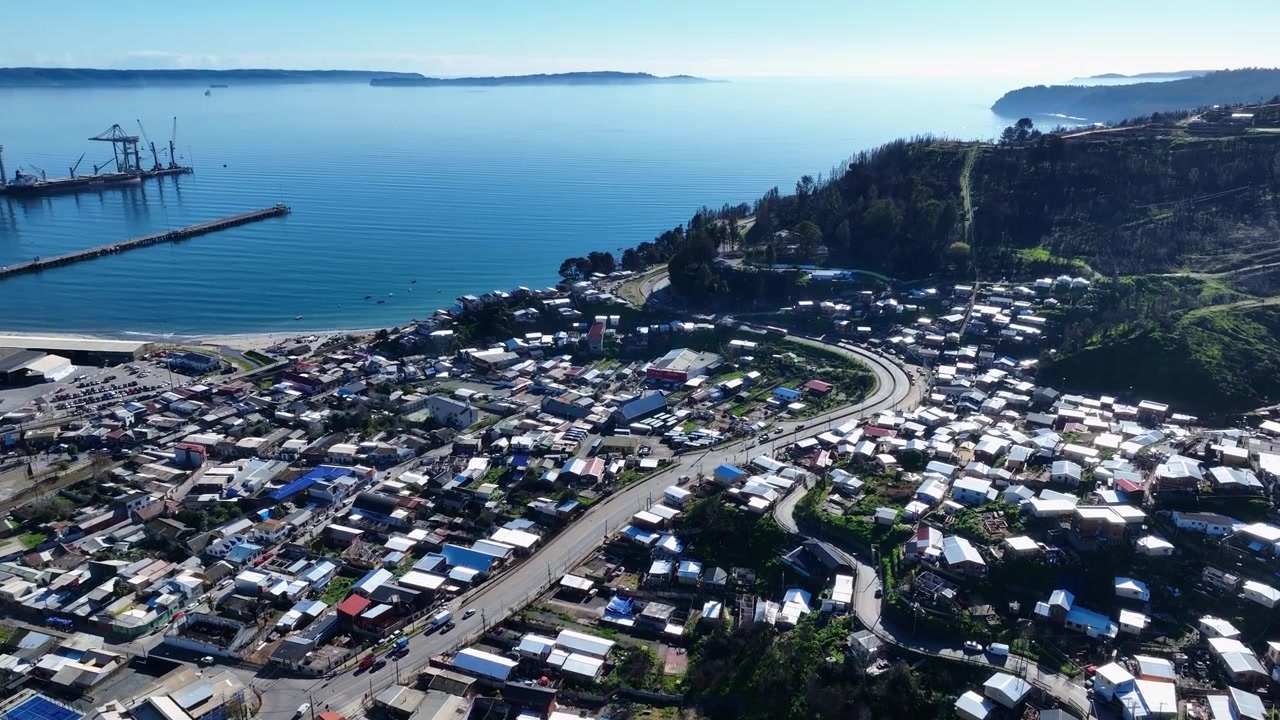Inquadratura dal basso verso l'alto che rivela una densa cittadina costiera annidata lungo strade tortuose e colline che si affacciano su una baia calma. Lirquen, Penco, Cile.