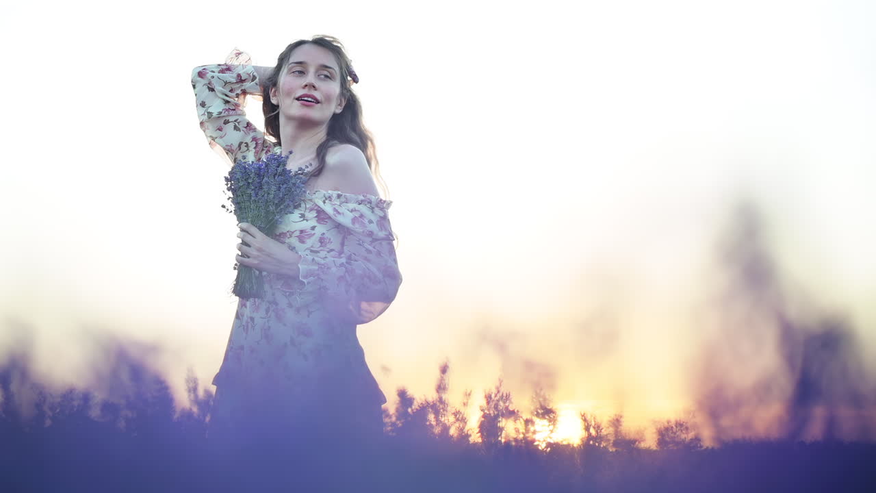 Young woman in floral dress holding a lavender bouquet and smiling warmly at golden hour