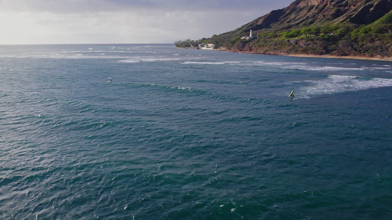 Riprese drone che si muovono verso la costa di Oahu mentre surfisti e kiteboarder punteggiano la superficie dell'oceano in una giornata di sole vicino a Diamond Head nelle Hawaii