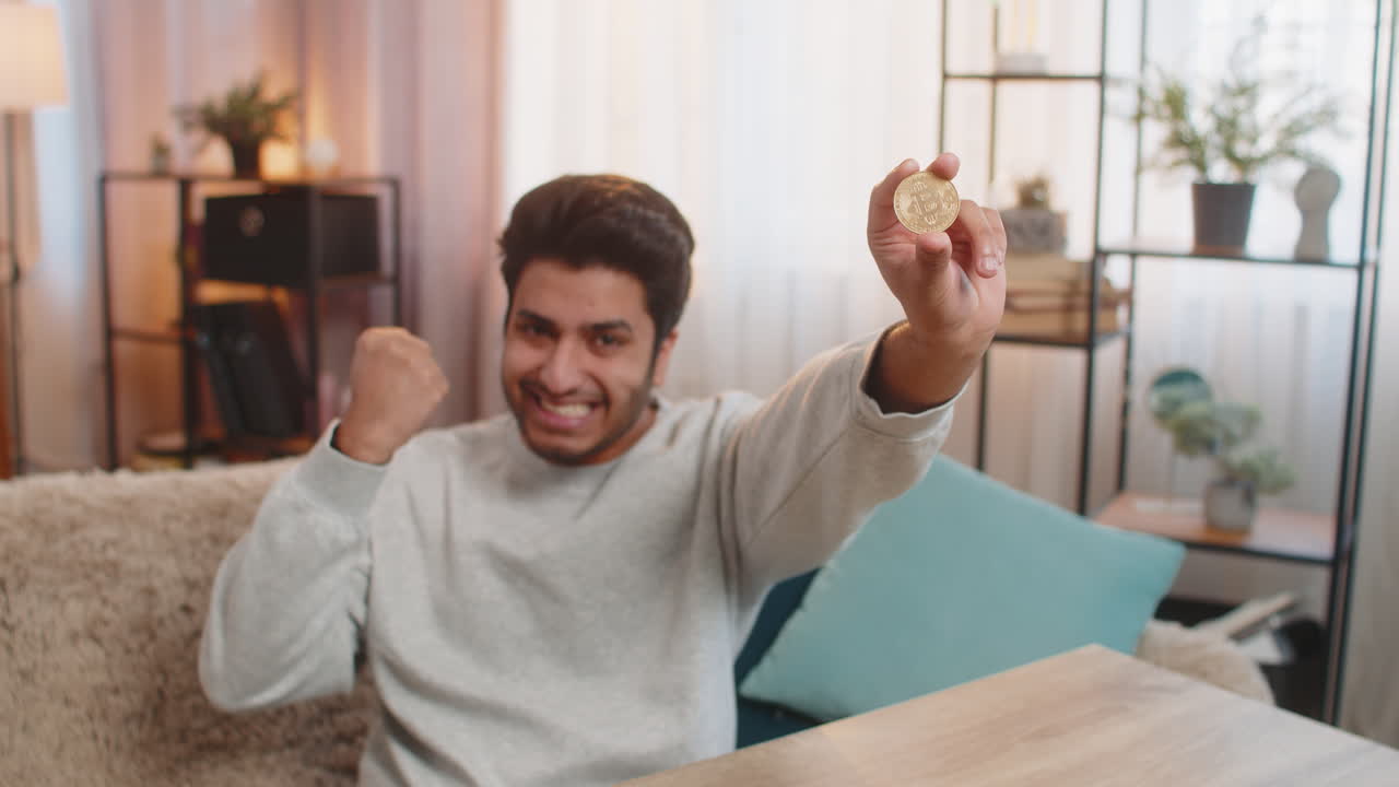 Joven indio celebrando que ha ganado criptomonedas y mostrando un bitcoin tras consultar los gráficos de cambio.