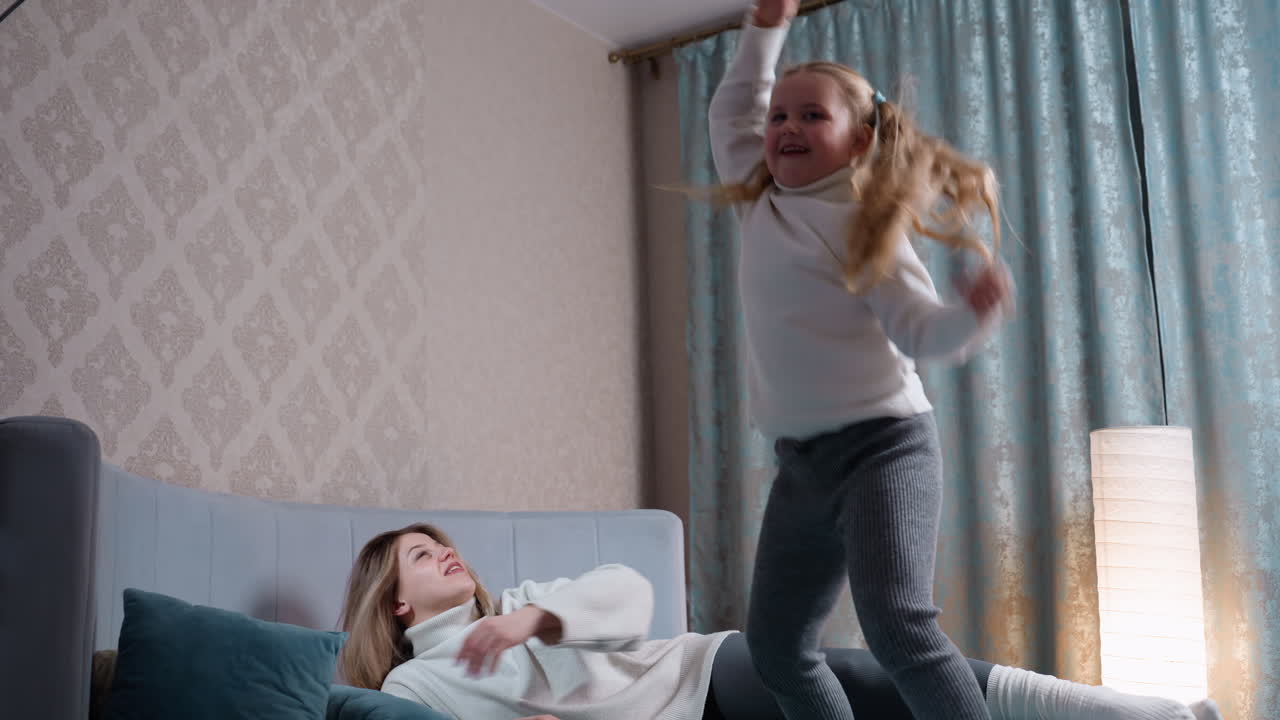 Cheerful mother relaxes on bed holding notebook while playful daughter jumps energetically nearby, creating dynamic scene of joy, fun, and family bonding in cozy bright bedroom with curtains