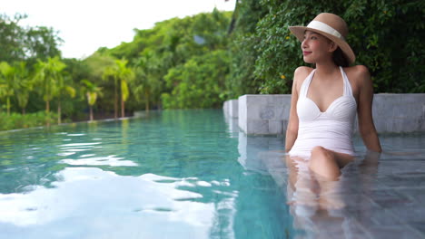 Pretty-Asian-woman-in-a-one-piece-white-bathing-suit-sits-on-the-shallow-step-of-a-resort-swimming-pool-looking-out-to-her-right