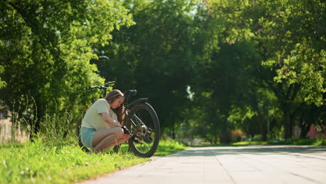 young female cyclist crouches on a sunlit grassy path, focused on pumping air into back tire of her bicycle, surrounded by lush greenery and trees