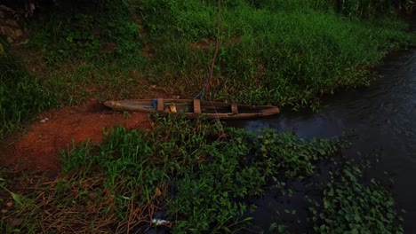 dolly in of a wooden canoe lying on the the riverbank of a calm river surrounded by tropical vegetation. filmed at eleyele river in ibadan, nigeria, africa